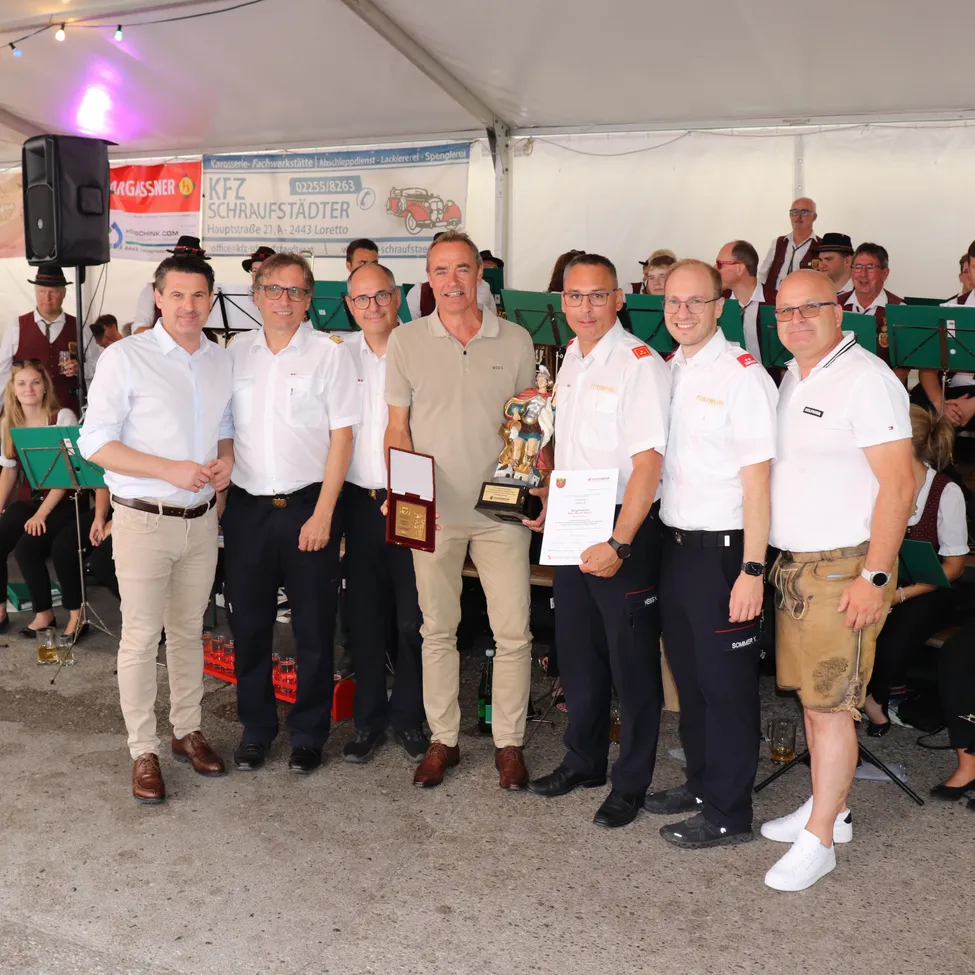 Eine Gruppe von Männern in Uniformen und Freizeitkleidung steht unter einem Zelt, hält einen Pokal und ein Zertifikat. Sie lächeln und posieren für ein Foto. Dahinter ist ein Banner mit Text und Logo zu sehen.