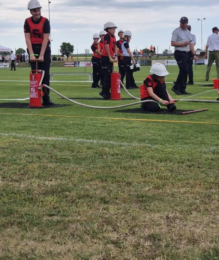 Eine Gruppe von Feuerwehrleuten in roten und weißen Uniformen trainiert auf einem Feld. Eine Person hält eine Feuerwehrschläuche, während eine andere auf dem Boden kniet. Im Hintergrund stehen Leute in der Nähe eines Zeltes.