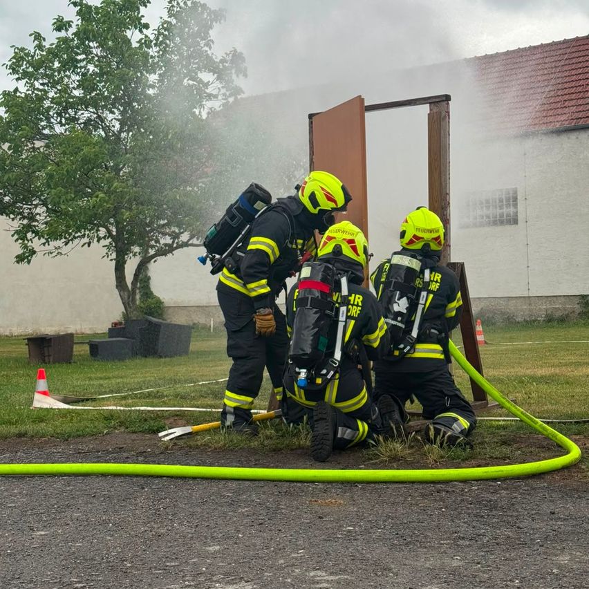 Three firefighters are outside in uniform, with one standing near a door and another spraying water. A green hose is laid on the ground.
