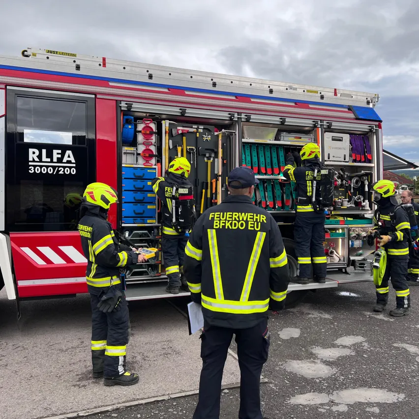 Eine Gruppe Feuerwehrleute steht um einen Feuerwehrwagen. Ein Mann in einem schwarzen Jackett mit gelbem Schriftzug sieht sich etwas an. Der Wagen hat ein Schild mit der Aufschrift RLFA 3000/200.