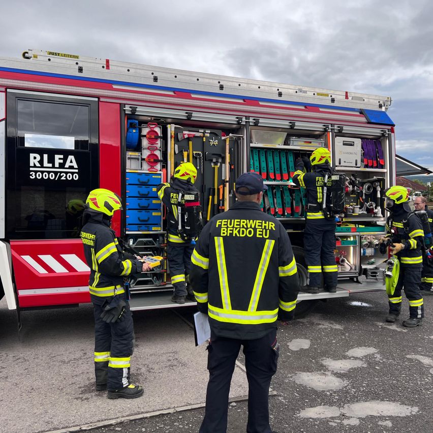A group of firefighters are gathered around a fire truck. A man in a black jacket with yellow writing is looking at something. The truck has a sign that reads RLFA 3000/200.