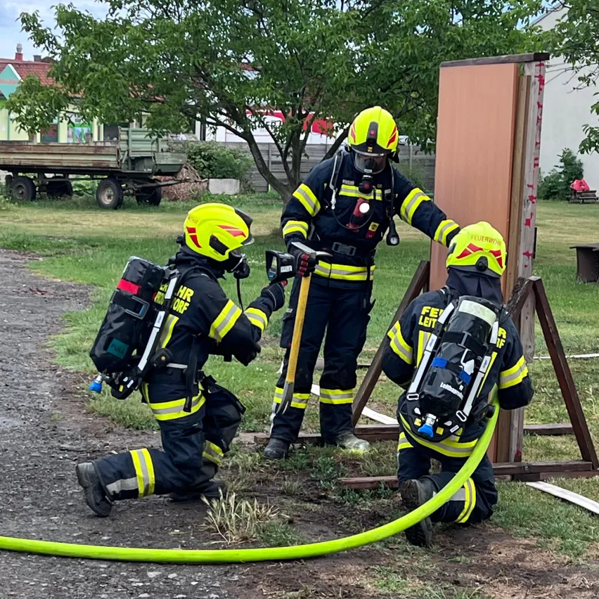 Drei Feuerwehrleute bereiten sich darauf vor, ein Feuer zu löschen. Einer kniet, ein anderer steht hinter einer Struktur und der dritte hält einen Schlauch. Bäume und ein Gebäude im Hintergrund.