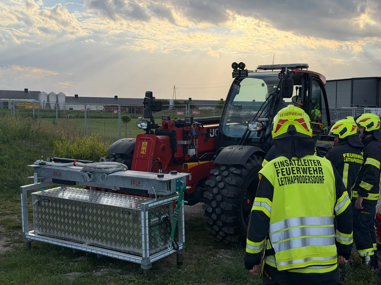 Ein Feuerwehrmann steht neben einem großen roten Traktor mit einer Metall-Werkzeugkiste und einem angeschlossenen Schlauch, unter einem teilweise bewölkten Himmel.