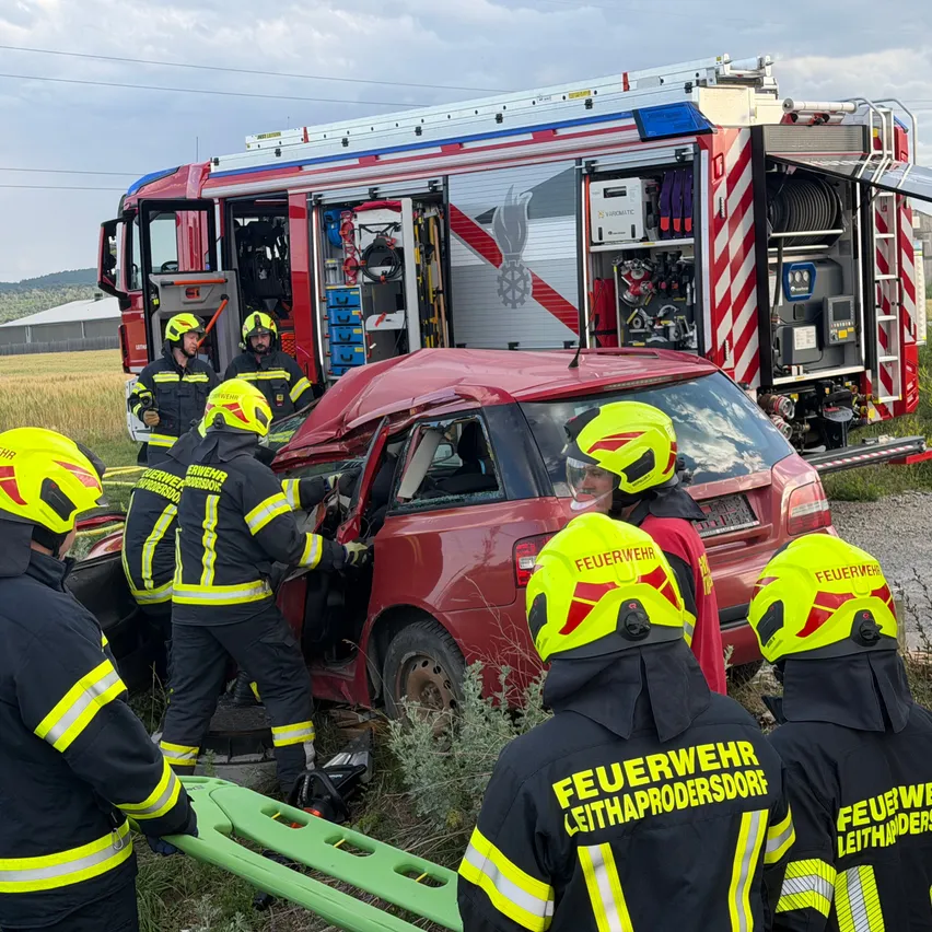 Feuerwehrleute in Schutzausrüstung helfen einem roten Auto mit zerbrochenem Fenster und Tür, das auf einem Rasen steht, mit einem Feuerwehrauto im Hintergrund.