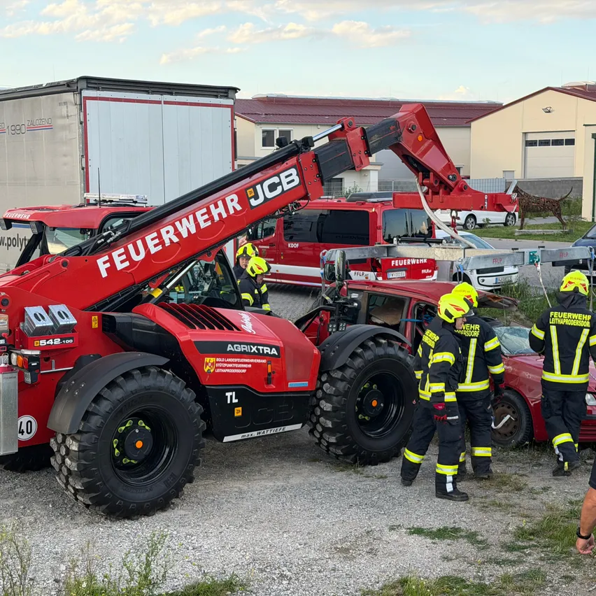 Feuerwehrleute in gelben Helmen verwenden einen roten JCB AgriXtra-Fahrzeug, um ein beschädigtes rotes Auto anzuheben. Die Szene spielt sich in einem offenen Bereich ab, mit Gebäuden und geparkten Fahrzeugen im Hintergrund.