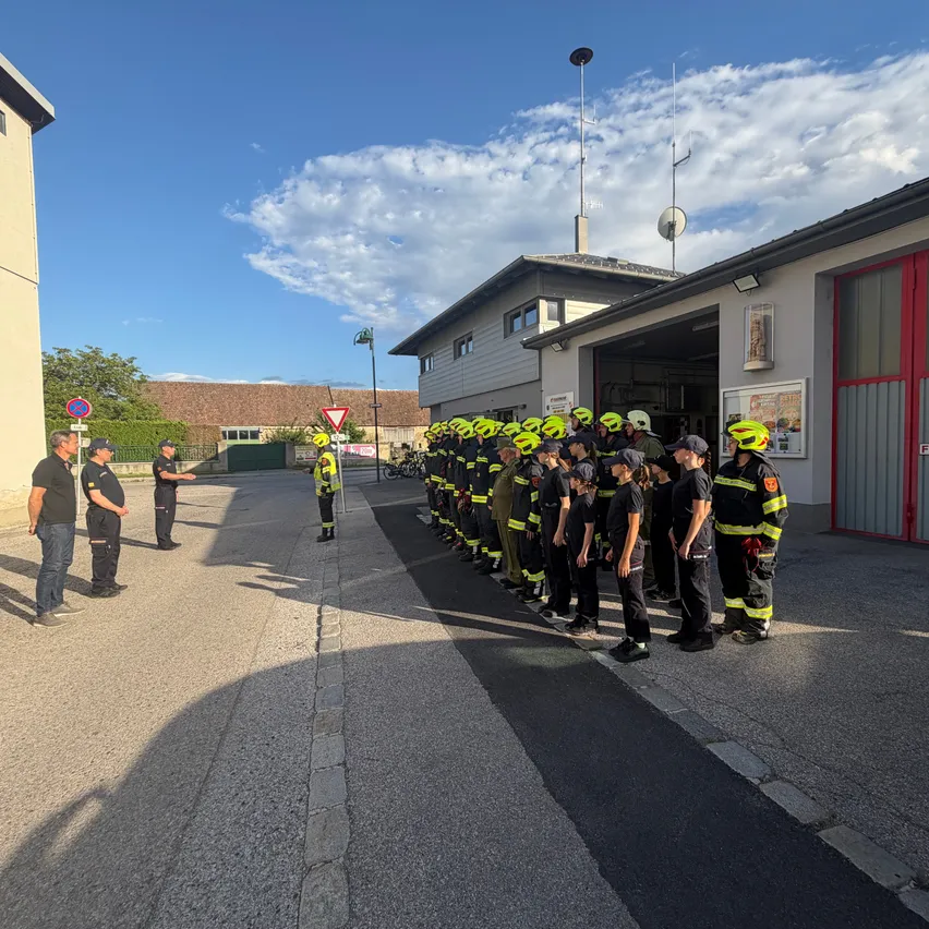 Eine Reihe von Feuerwehrleuten in Uniform steht vor einem Feuerwehrhaus unter einem blauen Himmel. Zwei Personen befinden sich hinter der Reihe.