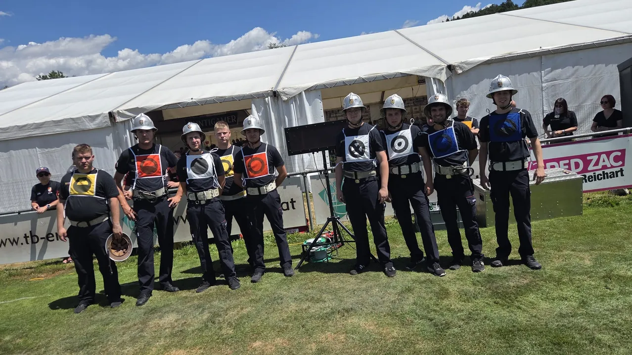 Eine Gruppe junger Männer in Uniformen steht in einer Reihe auf einem Rasenfeld, trägt Helme und Gürtel mit verschiedenen Abzeichen. Hinter ihnen steht ein großes Zelt mit einem Banner, und ein Lautsprecher ist davor aufgestellt.