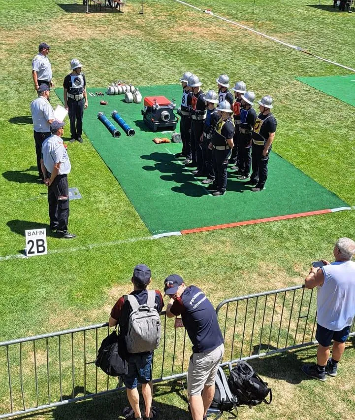 Gruppe uniformierter Personen mit Helmen steht auf einem markierten Feld, mit Offiziellen in der Nähe. Zuschauer beobachten von einer Absperrung aus. Ausrüstung ist auf dem Feld sichtbar.
