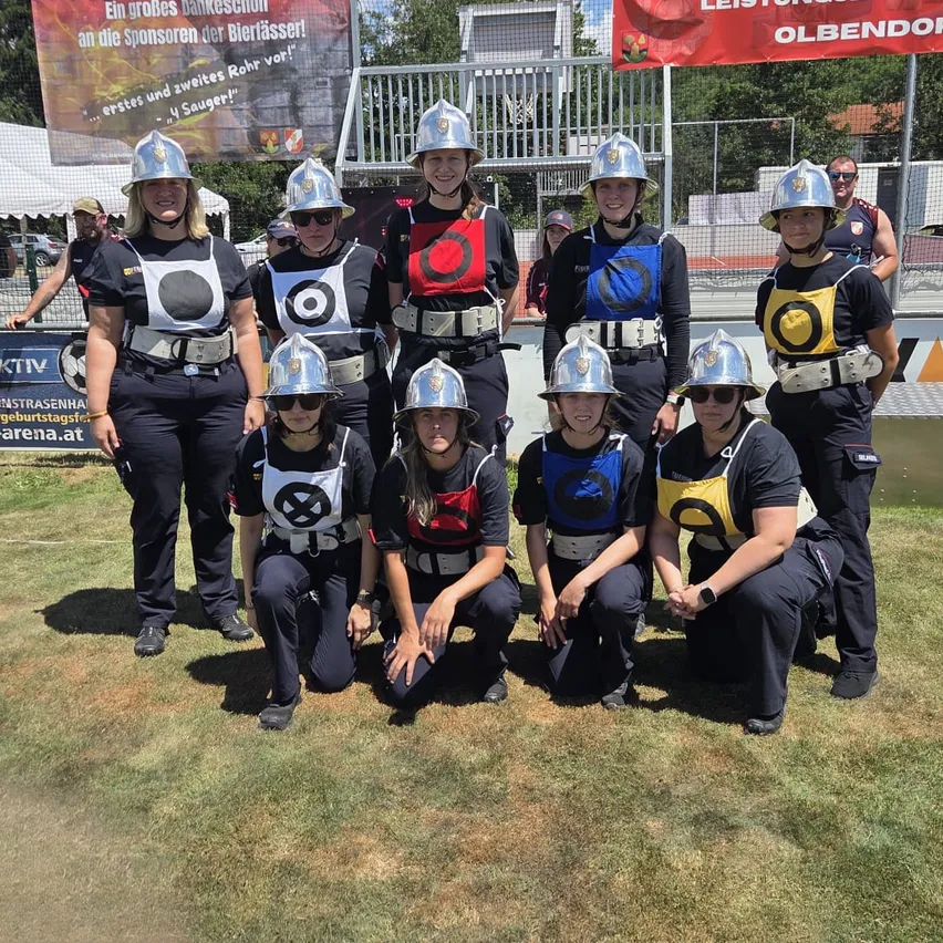 Eine Gruppe von Frauen in gleichen Uniformen und Helmen posiert für ein Foto auf einem Feld. Dahinter ist ein Zaun und ein Banner mit deutschem Text zu sehen.