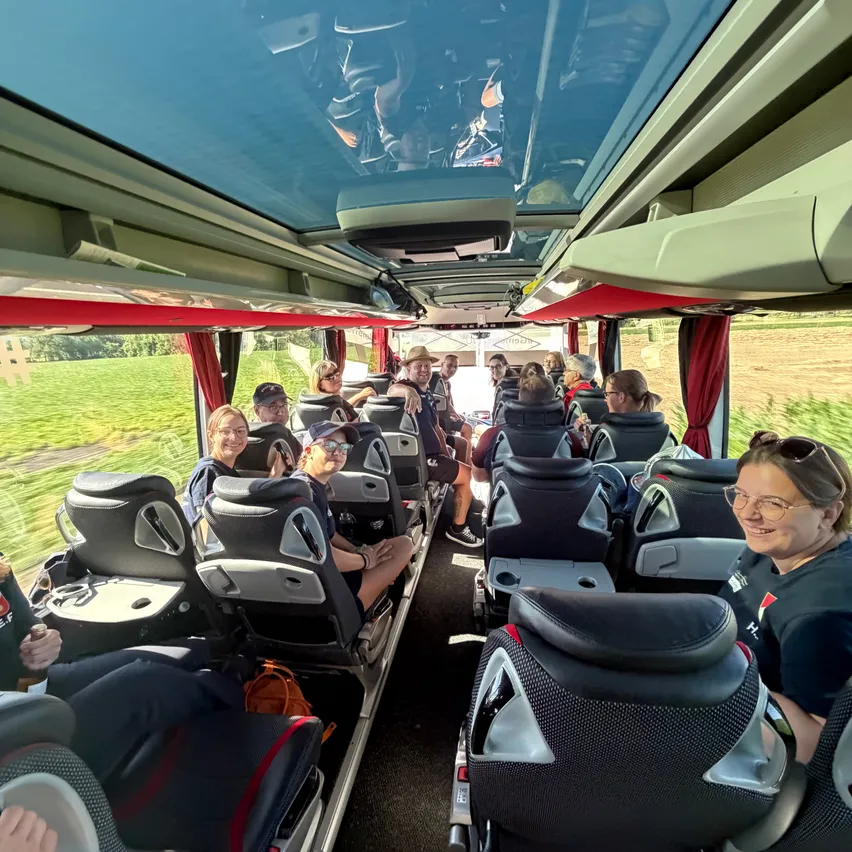Ein Bus fährt mit mehreren Passagieren auf einer Straße, einige tragen Hüte und Brillen. Die Sitze sind dunkelgrau mit roten Besätzen, und das Innere wird durch Sonnenlicht beleuchtet.