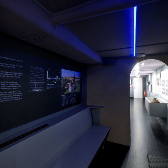 Eine Museumsausstellung zeigt einen großen Bildschirm mit Text und einem Bild einer Kirche, begleitet von einer Bank und einer blau beleuchteten Decke. Der Raum ist schwach beleuchtet, und ein kleiner Regal mit einer Pflanze ist sichtbar.