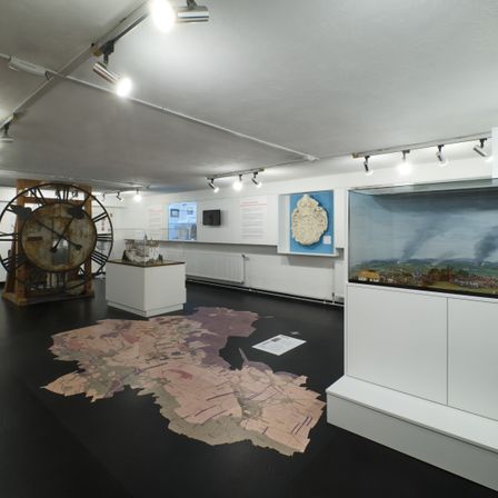 Ein Raum mit einer großen Uhr, einer Karte auf dem Boden und einem Landschaftsgemälde in einem Glaskasten. Es gibt auch weiße Schränke und ein kleines Fenster.
