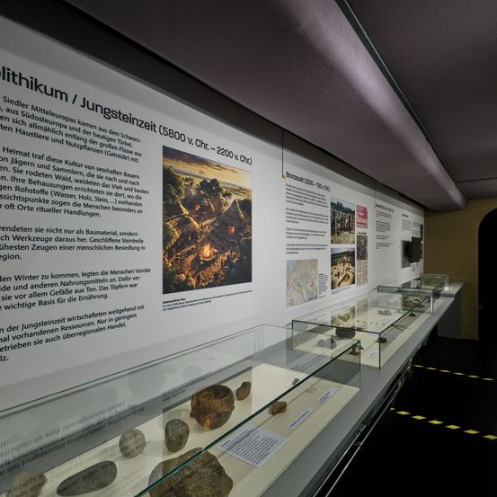 Eine Museumsausstellung zeigt jungsteinzeitliche Werkzeuge und Artefakte mit einem Informationsbrett, das eine Illustration von jungsteinzeitlichen Siedlern enthält. Darunter befinden sich Vitrinen mit verschiedenen Steinen und Artefakten.