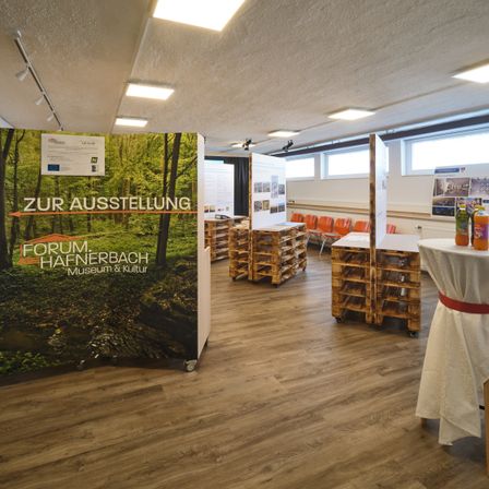 Ein Raum mit Holzböden, Deckenleuchten und einem Ausstellungsbanner. Orange Stühle und Tische sind aufgestellt, und ein Tisch hat Flaschen. Ein Schild steht 'Zur Ausstellung'.