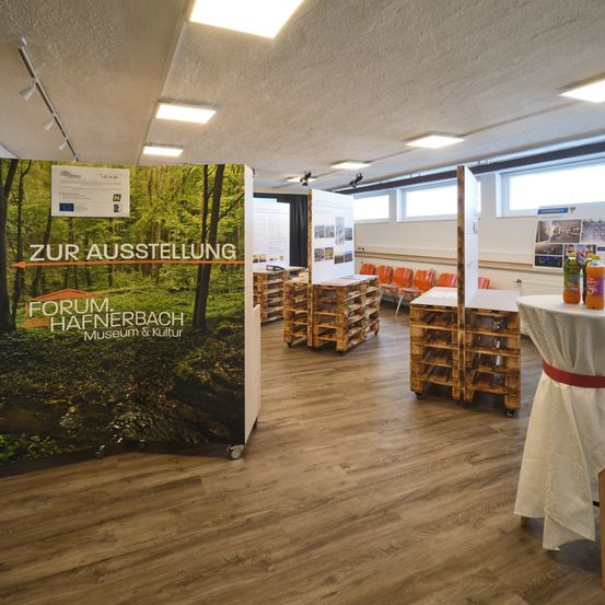 Ein Raum mit Holzböden, Deckenleuchten und einem Ausstellungsbanner. Orange Stühle und Tische sind aufgestellt, und ein Tisch hat Flaschen. Ein Schild steht 'Zur Ausstellung'.