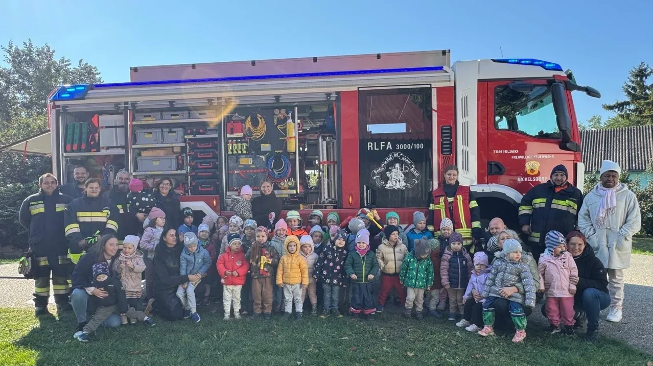 Eine Gruppe von Kindern und Erwachsenen posiert vor einem Feuerwehrauto mit dem Label RLFAA 3000/100. Das Auto steht auf einem Rasen.