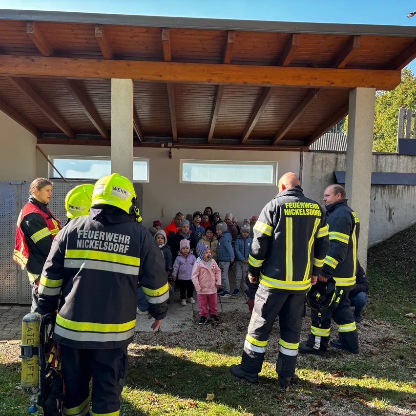 Feuerwehrleute aus Nickelsdorf stehen mit Kindern in einem geschützten Bereich. Die Feuerwehrleute tragen Uniformen mit reflektierenden gelben und schwarzen Streifen. Die Kinder sind versammelt und lauschen den Feuerwehrleuten.