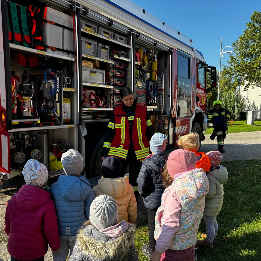 Eine Gruppe von Kindern steht vor einem Feuerwehrauto und hört einem Feuerwehrmann zu, der ihnen die Ausrüstung zeigt.