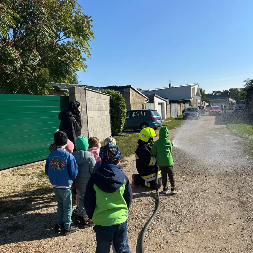 Kinder und Erwachsene beobachten einen Feuerwehrmann, der Wasser auf die Straße sprüht, in der Nähe eines grünen Tores und Gebäude.