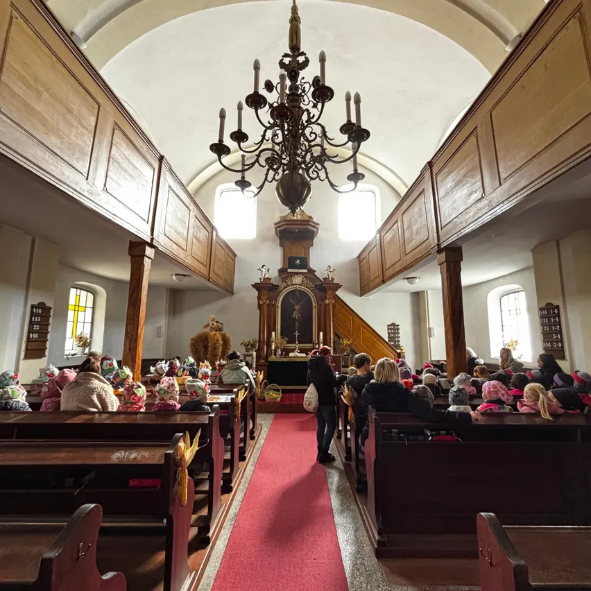 Ein Kircheninneres mit Holzbänken, einem roten Teppich und einem Kronleuchter. Menschen sitzen auf Bänken, einige tragen Mützen. Eine Person steht am Altar, und eine Statue ist auf dem Altar. Die Wände sind aus Holz, und es gibt Fenster mit Buntglas.