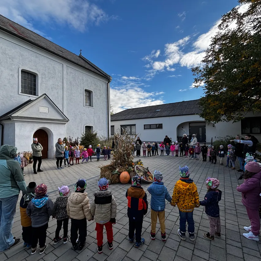 Eine Gruppe von Kindern in Winterkleidung bildet einen Kreis vor einer Kirche an einem sonnigen Tag. Einige Erwachsene stehen in der Nähe.