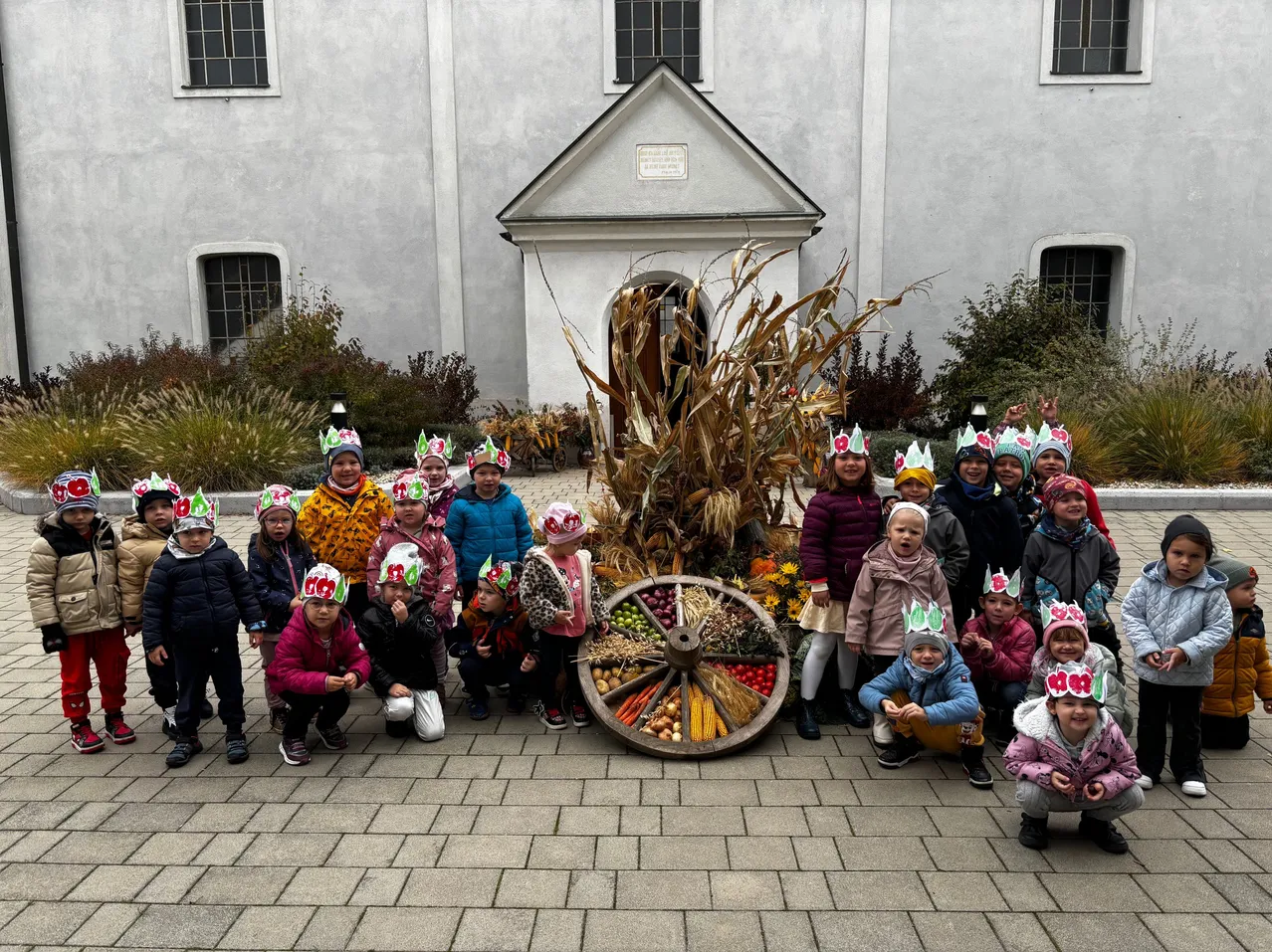 Eine Gruppe von Kindern mit festlichen Mützen posiert für ein Foto vor einem dekorativen Rad und einem Maiskolben, mit einer Kirche im Hintergrund.
