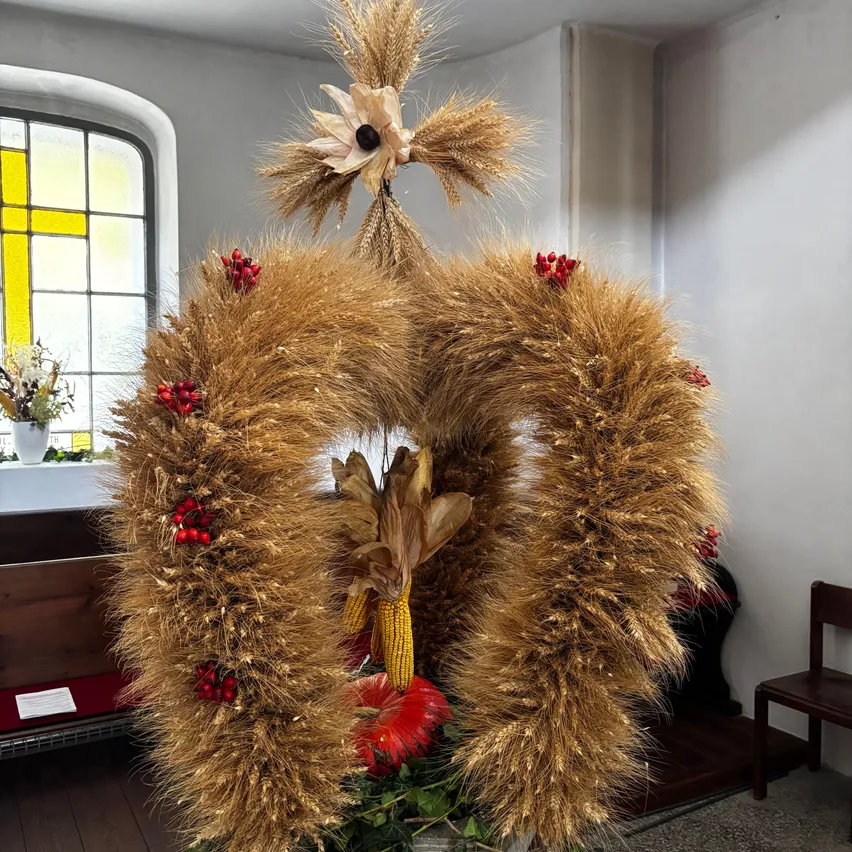 Ein goldener Weizenkranz mit roten Beeren hängt an einer weißen Wand. Ein Holzstuhl und ein Holztisch mit einer weißen Vase sind im Hintergrund.