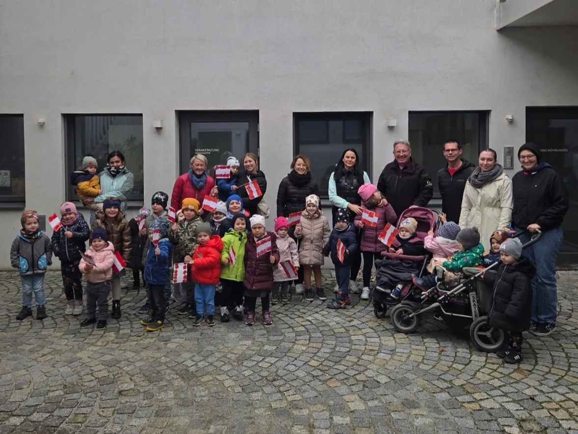Eine Gruppe von Kindern, Erwachsenen und Eltern versammelt sich vor einem Gebäude, alle halten Flaggen. Einige Erwachsene tragen Brillen und einige Kinder tragen Mützen. Ein Kinderwagen ist vorhanden.