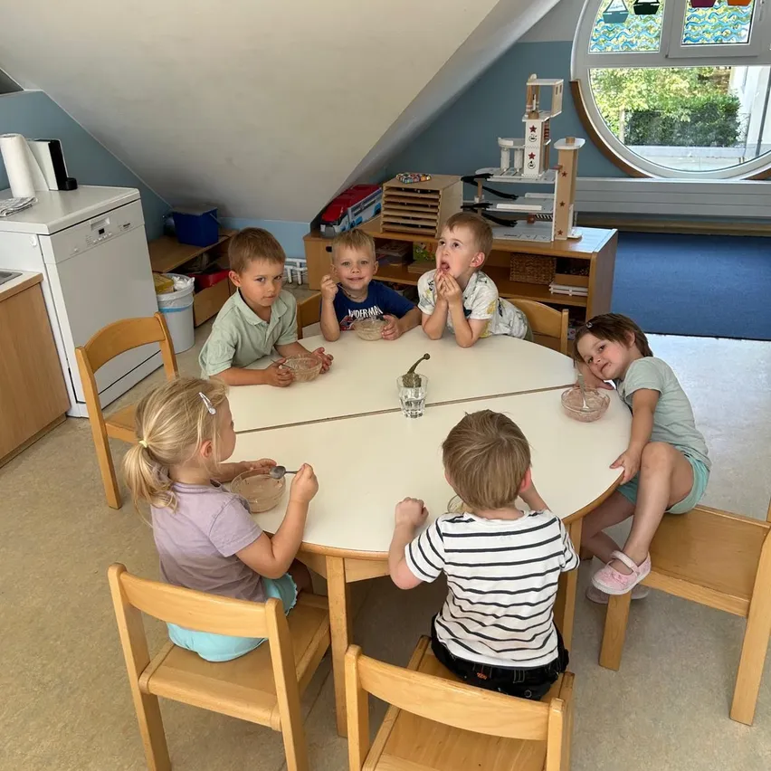 Eine Gruppe junger Kinder sitzt um einen runden Tisch herum, jeder hält eine Schüssel und einen Löffel, möglicherweise essen oder bereiten sie Essen zu. Der Raum hat Holzmöbel und ein großes kreisförmiges Fenster.
