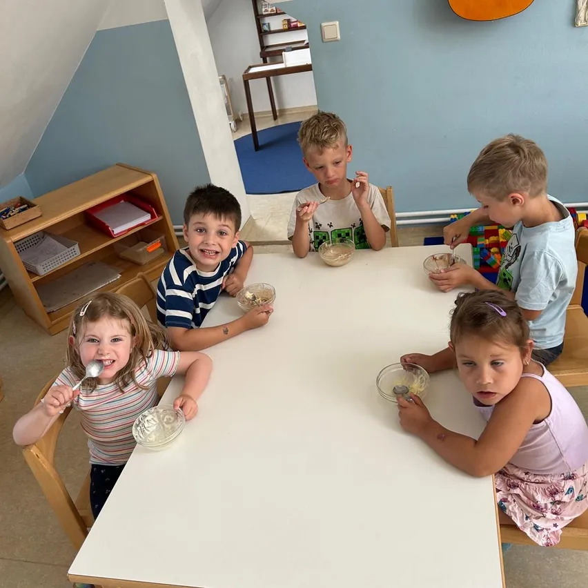 Eine Gruppe von Kindern, einschließlich Jungen und Mädchen, sitzt um einen Tisch herum, isst aus Schalen mit Löffeln und lächelt. Hinter ihnen steht ein Holzschrank und ein Teppich liegt auf dem Boden.
