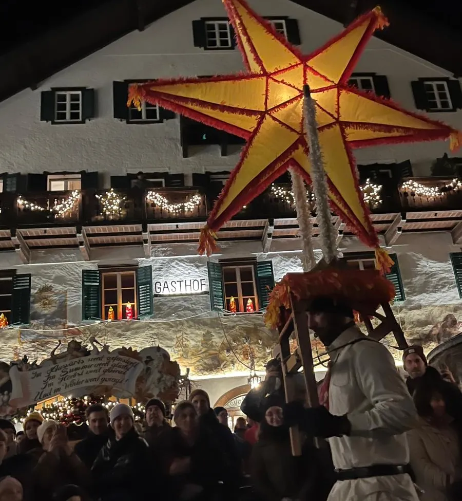 Ein Mann, der eine große Sternlaterne trägt, geht vor einem dekorierten Gebäude mit einem Banner. Viele Menschen schauen zu, und die Szene ist von Lichtern erhellt.