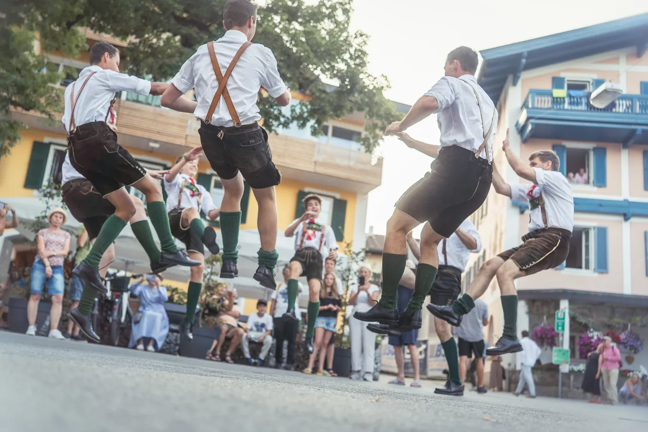 Eine Gruppe von Männern in traditioneller deutscher Kleidung führt einen lebhaften Tanz im Freien auf.