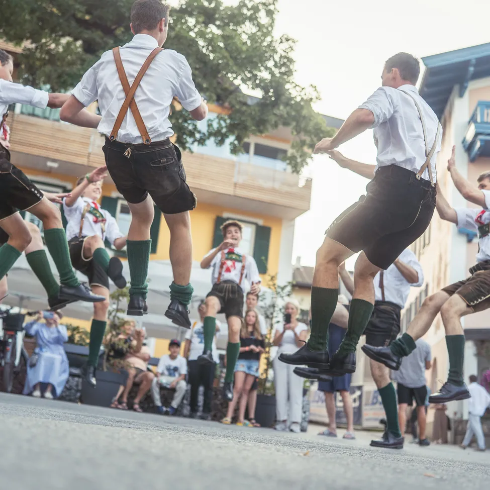 Eine Gruppe von Männern in traditioneller deutscher Kleidung führt einen lebhaften Tanz im Freien auf.