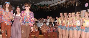 Eine Gruppe von Menschen in farbenfroher, retro Kleidung tritt auf einer Bühne auf. Ein Mann in einem Tie-Dye-Shirt und Glockenhosen geht vor ihnen.