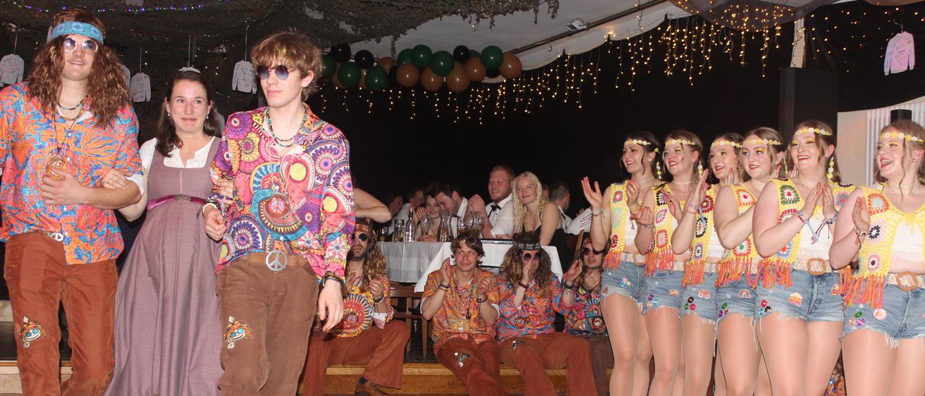 Eine Gruppe von Menschen in farbenfroher, retro Kleidung tritt auf einer Bühne auf. Ein Mann in einem Tie-Dye-Shirt und Glockenhosen geht vor ihnen.