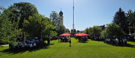 Eine Gruppe von Menschen in traditioneller Kleidung versammelt sich auf einem Rasenfeld unter roten Sonnenschirmen, mit einem Kirchturm im Hintergrund.