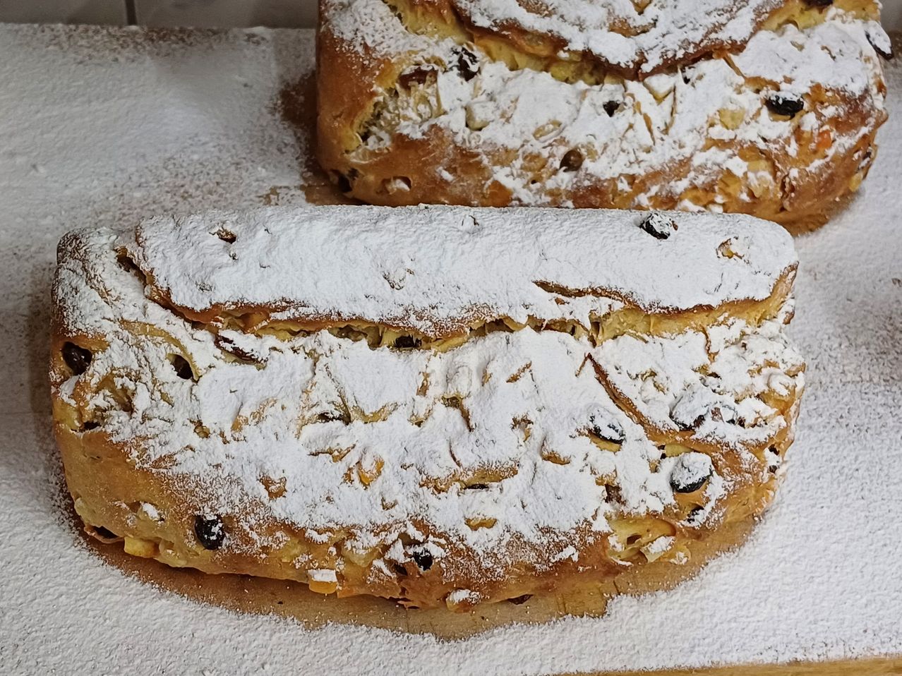 Zwei Brote liegen auf einer weißen Oberfläche, bestreut mit Puderzucker und gefüllt mit Rosinen und Nüssen.