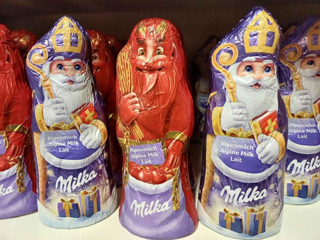 Drei Schokoladenfiguren von Santa Claus sind ausgestellt, jede in einer anderen Farbe. Sie halten ein Buch und einen roten Beutel, und der Name 'Milka' ist auf ihrer Verpackung deutlich sichtbar.