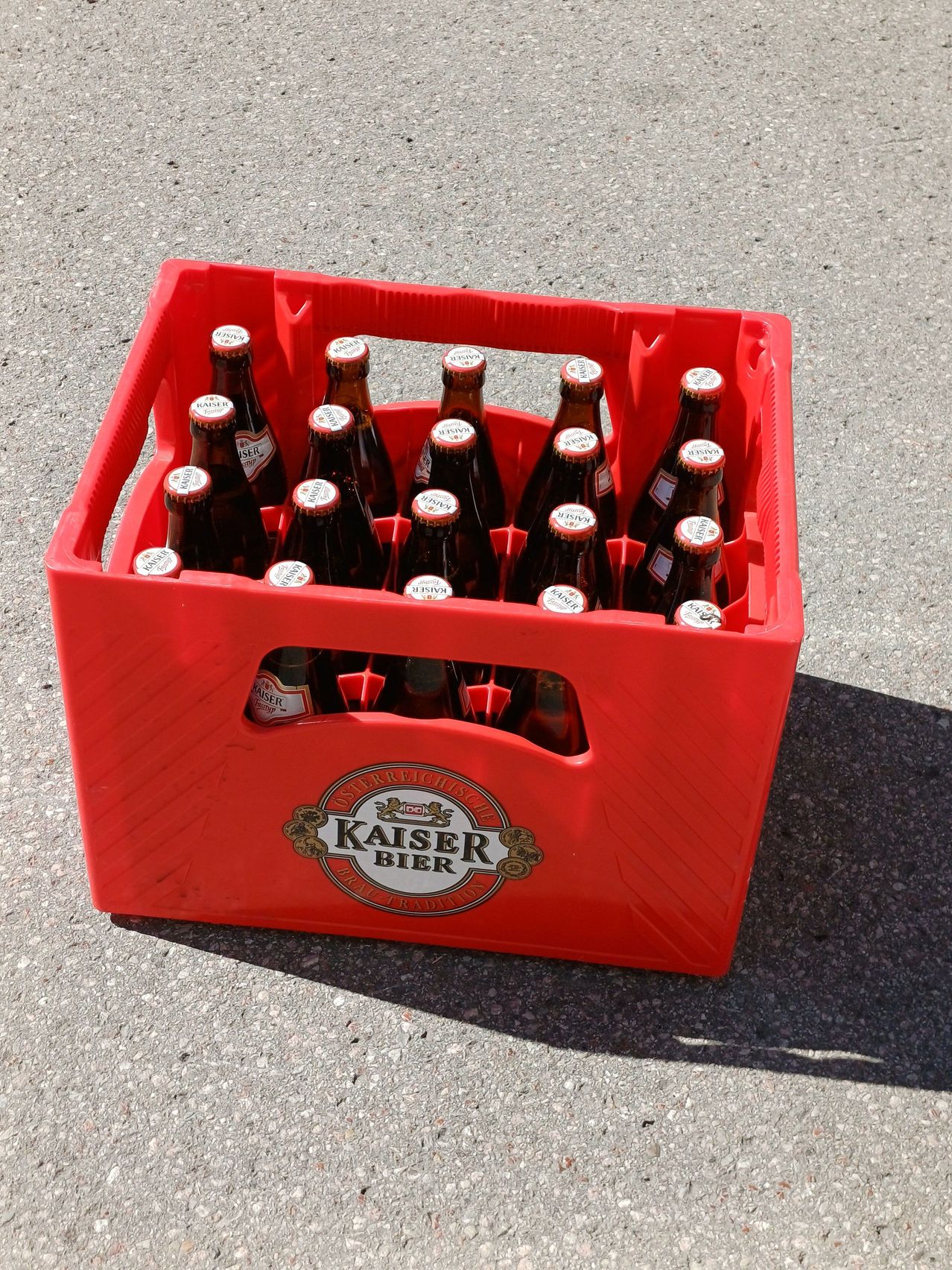 Ein roter Plastikkasten gefüllt mit Flaschen von Kaiser Bier, einer deutschen Biermarke, auf einer Betonoberfläche.