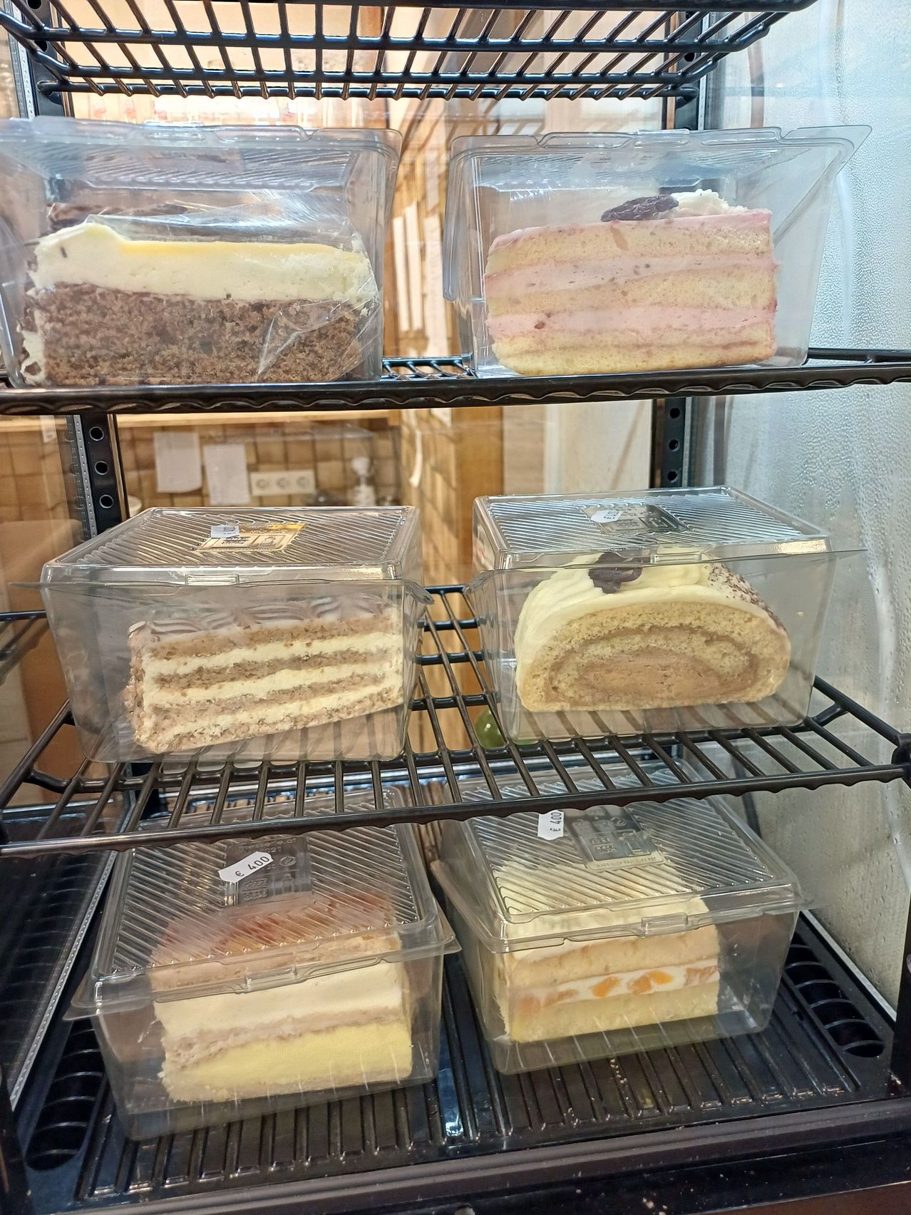Eine Vitrine mit verschiedenen Kuchen in durchsichtigen Plastikbehältern, darunter ein Schokoladenkuchen, ein rosa gefrorener Kuchen, ein mehrschichtiger Kuchen und ein Rollkuchen.