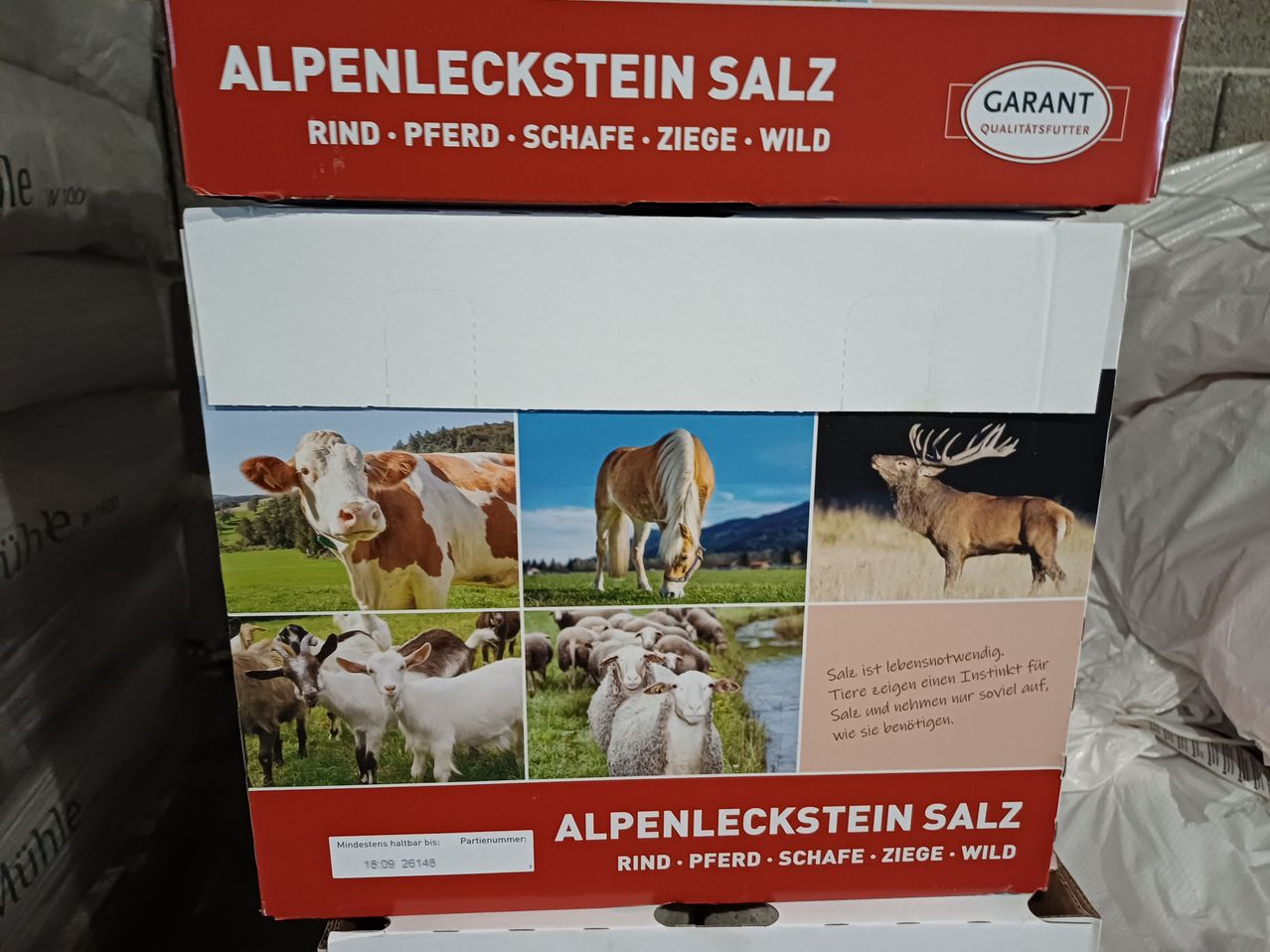 Eine Schachtel Alpenleckstein Salz, ein Qualitätsfutter für Rinder, Pferde, Schafe, Ziegen und Wildtiere, mit Bildern verschiedener Tiere. Die Schachtel hat ein rot-weißes Etikett mit dem Markennamen 'GARANT' und dem Produktnamen 'ALPENLECKSTEIN SALZ'.