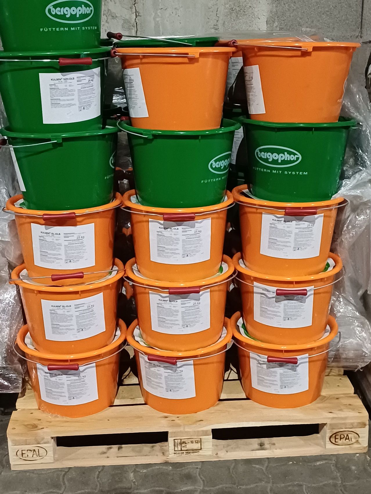 Ein Stapel orangefarbener und grüner Eimer mit weißen Etiketten, jeweils beschriftet mit 'Bergophor' und 'Futtermittel mit System', auf einem Holzpalette angeordnet.