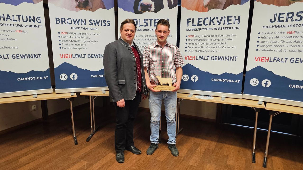 Zwei Männer stehen vor einem Hintergrund mit Nutztieren. Ein Mann hält einen Holzpreis. Der Hintergrund zeigt Kühe und die Texte "Holstein, Mehr als Milch" und "Fleckvieh, Doppelnutzung in Perfektion."