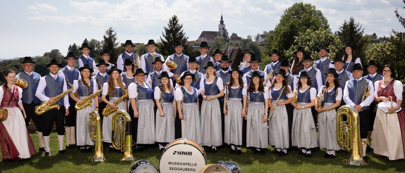 Ein Musikband in traditioneller Kleidung posiert für ein Foto an einem sonnigen Tag mit Instrumenten wie Saxophonen, Trompeten und Trommeln im Vordergrund.