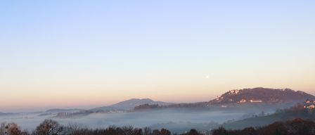 Eine neblige Landschaft mit Bergen und einem kleinen Dorf in der Ferne. Der Himmel ist klar mit einem Hauch von Orange am Horizont.