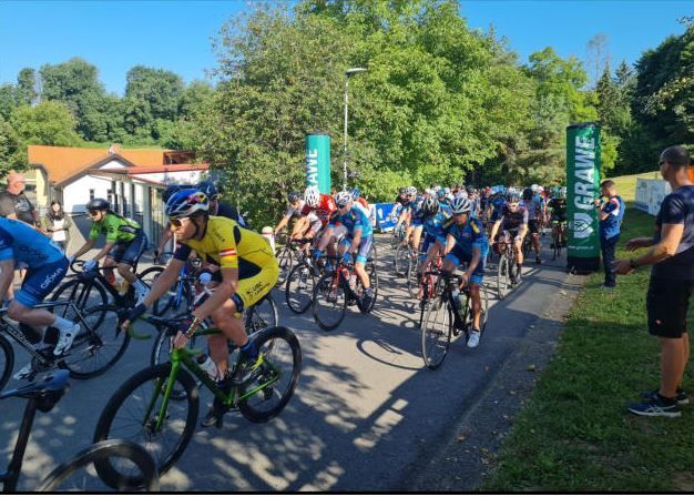 Eine Gruppe von Radfahrern nimmt an einem Rennen auf einer Straße teil, mit Bäumen und einem Gebäude im Hintergrund.