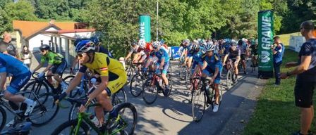 Eine Gruppe von Radfahrern nimmt an einem Rennen auf einer Straße teil, mit Bäumen und einem Gebäude im Hintergrund.