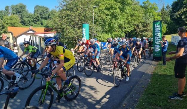 Eine Gruppe von Radfahrern nimmt an einem Rennen auf einer Straße teil, mit Bäumen und einem Gebäude im Hintergrund.
