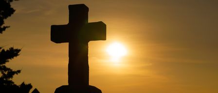 Eine Silhouette eines Kreuzes auf einem Hügel, die Sonne geht hinter ihm unter und schafft eine ruhige und spirituelle Atmosphäre.