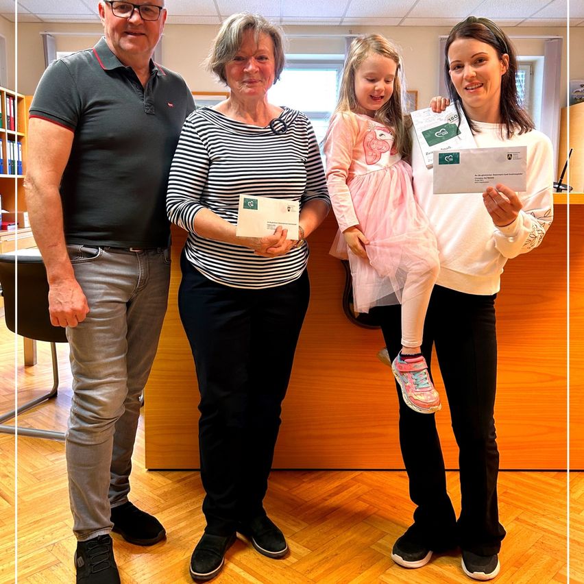 Vier Personen stehen in einem Büro und halten Urkunden in den Händen. Ein Mann und eine Frau in Schwarz-Weiß-Streifen, ein Mädchen in Pink und eine Frau in Weiß. Hinter ihnen steht ein Holzschreibtisch, Bücherregale und ein Stuhl.